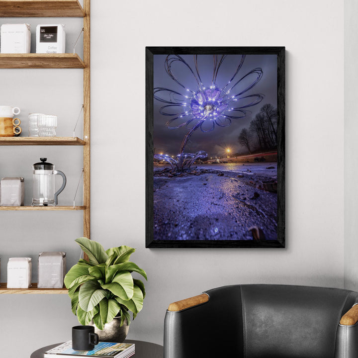 A tall wire-sculpted flower illuminated by small purple LED lights stands on wet pavement at night, with streetlights and a dramatic cloudy sky in the background, creating a surreal and atmospheric glowing flower art scene.